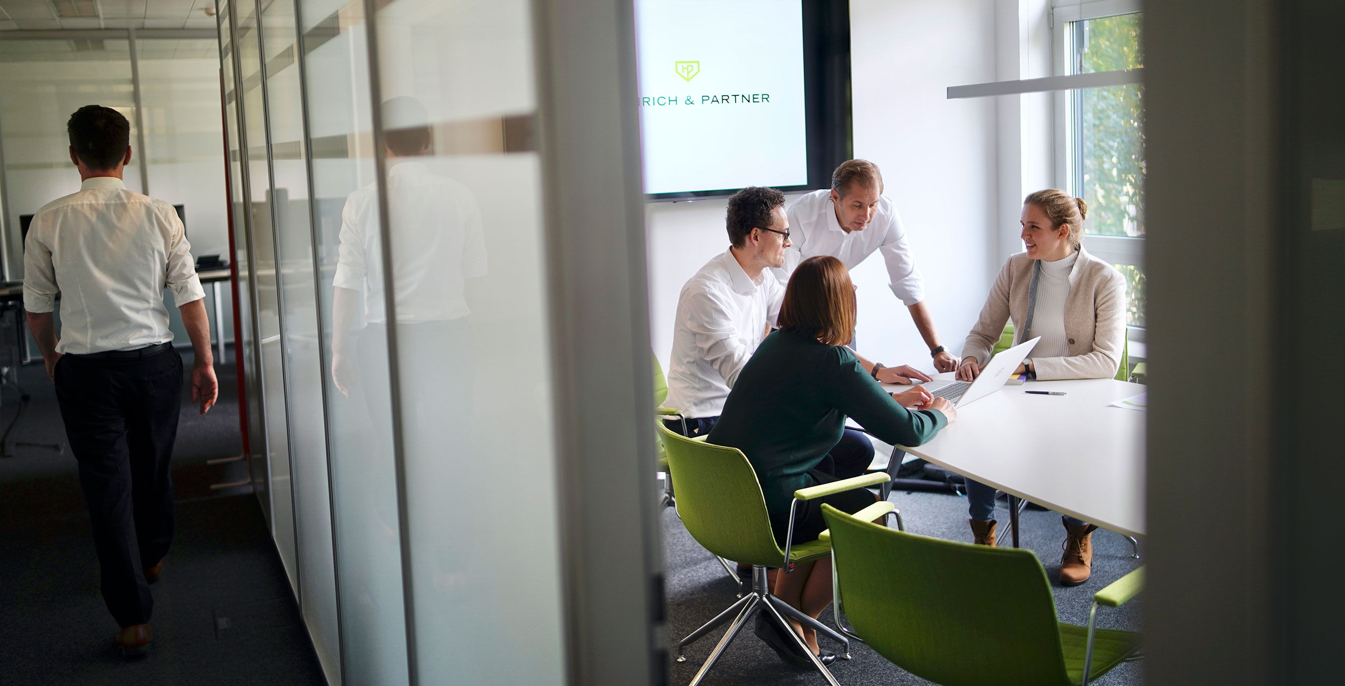 Eine Gruppe von fünf Personen sitzt in einem modernen Büro an einem Tisch. Vier Personen sind in ein Gespräch vertieft, während eine Person steht. Ein Mann geht im Hintergrund am Glaskorridor vorbei.